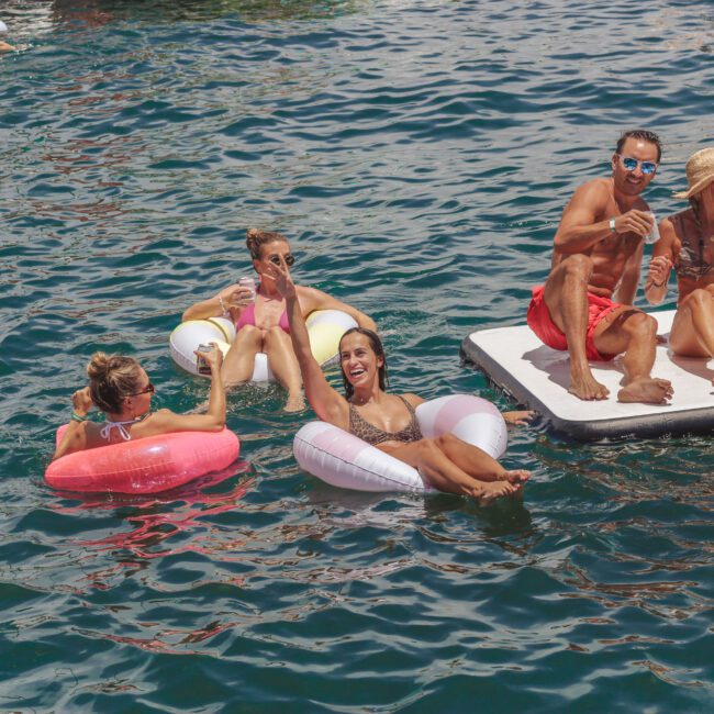 People relax and smile on colorful floaties and a floating platform in a sunny, vibrant blue body of water, enjoying a social event together.