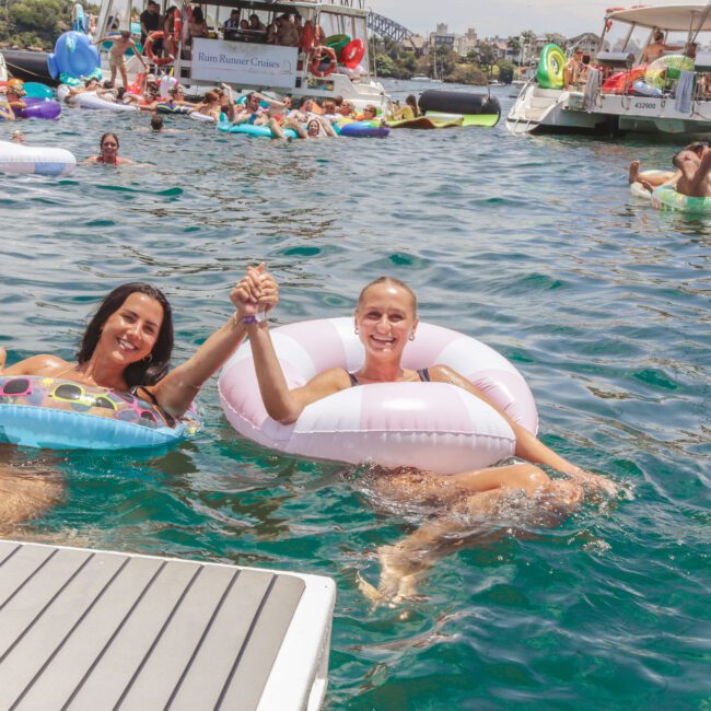 Two women in colorful pool floats smile and hold hands while relaxing in the water at a lively boat party, surrounded by other people, floats, and boats on a sunny day.