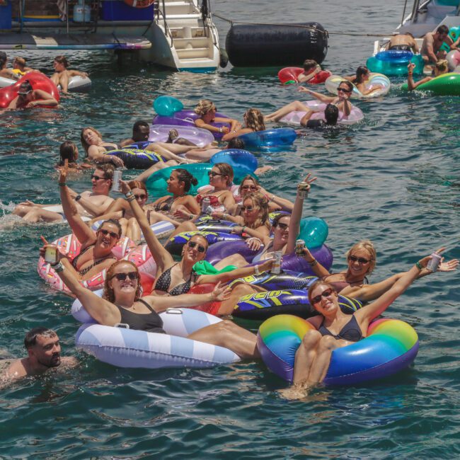 A group of people relax on colorful inflatable floats in the water near boats, smiling and waving, enjoying a sunny day at a lively social event.
