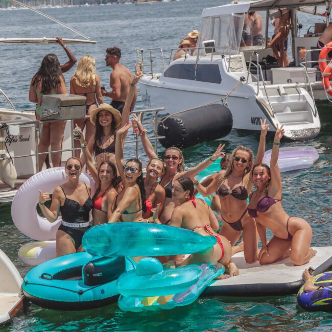 A group of people in swimsuits pose and smile on a floating platform with inflatable pool floats near docked boats on a sunny day. Others relax and socialize on nearby yachts in the background.