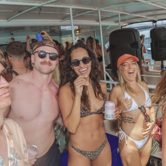 A group of young adults in swimwear smile and pose together on a boat during a lively daytime party. People in the background are also enjoying the festive atmosphere.