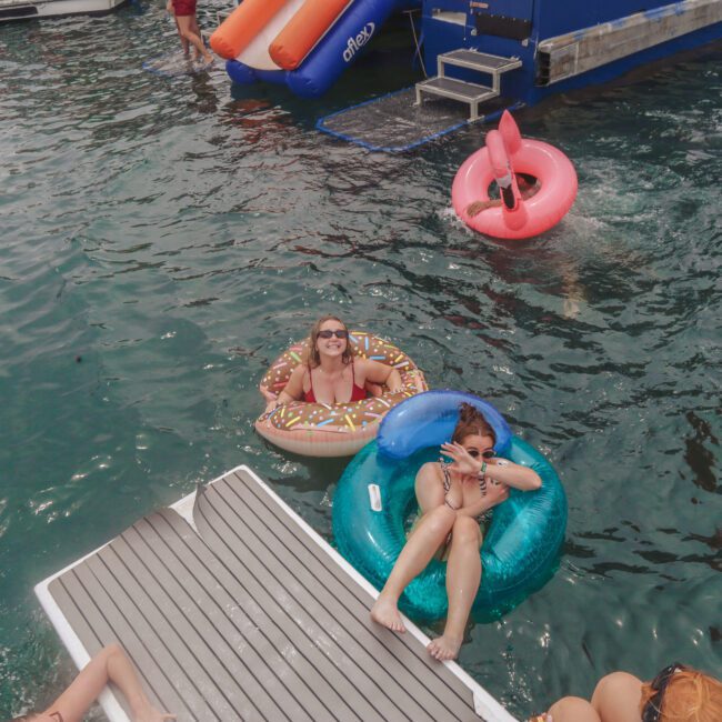 Two people in pool floats, one shaped like a donut and the other round and blue, smile and gesture at the camera near a docked boat. Other floats and a water slide are visible in the background.
