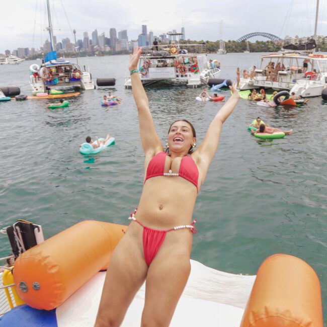 A woman in a red bikini slides down an inflatable water slide on a boat, arms raised and smiling, with people on boats and inflatables in the water, and a city skyline and bridge in the background.