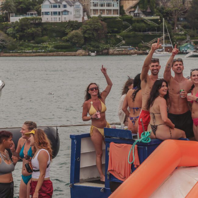 A group of young adults in swimwear smile, dance, and enjoy drinks on a boat, with water and waterfront houses in the background. Some are on an orange slide, while others gather and talk nearby.