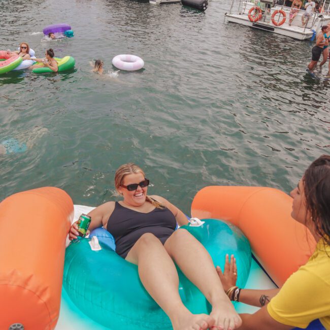 People relax on colorful pool floats in a lake near boats. A woman in sunglasses lounges on an inflatable with a drink, smiling and talking to another woman nearby. Others swim and float on the water in the background.