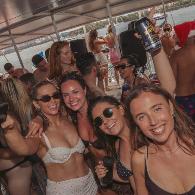 A large group of young adults in swimsuits smile, dance, and hold drinks on a crowded boat party under a canopy, with water and shoreline visible in the background.