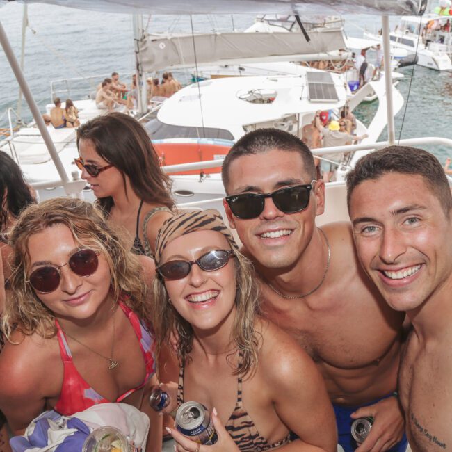 A group of young adults in swimsuits smile for a photo on a crowded boat party, with others mingling and yachts visible on the water in the background. The atmosphere is lively and sunny.