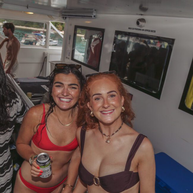 Two women in swimsuits smiling at the camera on a boat, holding drinks. Other people are in the background, some also in swimwear, with sunlight coming in from outside. The scene appears festive and social.