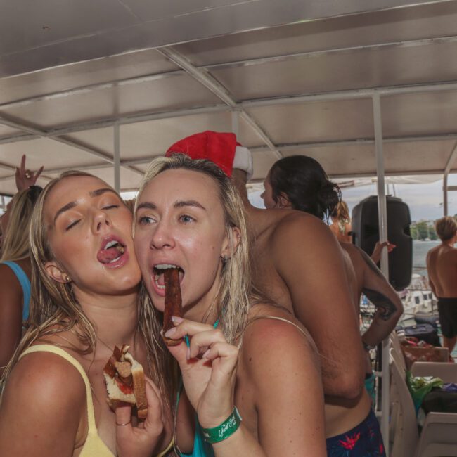Two women in swimsuits pose playfully on a crowded boat, one eating a sandwich and the other licking an ice pop. Others in swimwear party in the background under a canopy, suggesting a lively summer boat party.