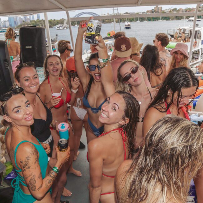 A group of young women in swimsuits smile and pose for the camera on a crowded boat during a lively party, holding drinks with water and more boats visible in the background.