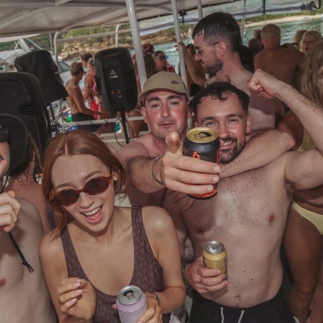 A group of young adults in swimwear enjoy drinks and smile for the camera at a lively boat party, with water and greenery visible in the background. The atmosphere is festive and energetic.