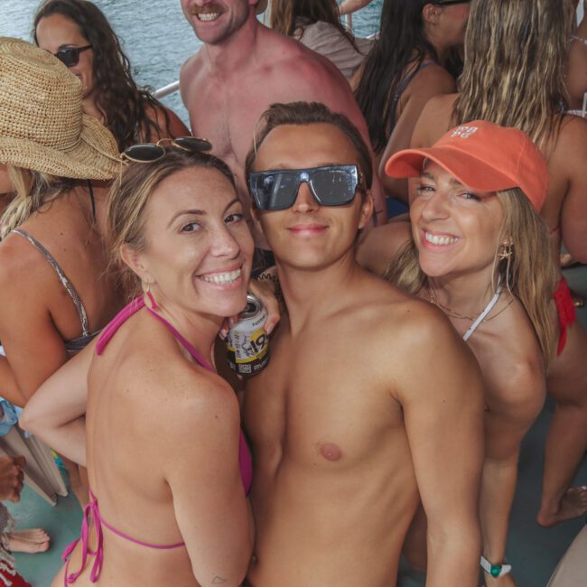 A group of young adults in swimsuits smile and pose together on a boat, holding drinks and enjoying a lively party by the water.