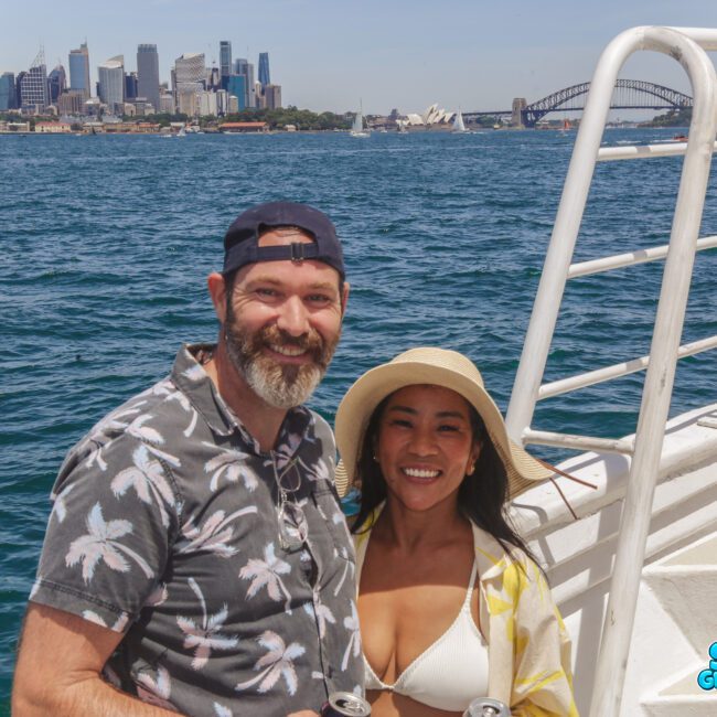A smiling man and woman pose on a boat with blue water and the Sydney skyline, including the Opera House and Harbour Bridge, in the background. The man wears a floral shirt and cap; the woman wears a sunhat and white cover-up.