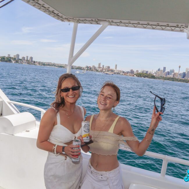 Two women in light summer outfits smile and pose on a boat, holding drinks with Sydney's city skyline and blue water in the background under a clear sky.