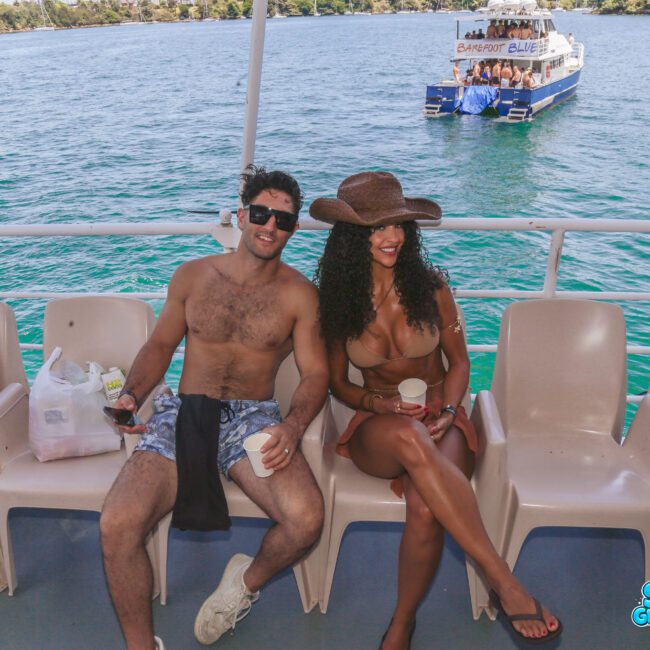 A man and woman in swimwear sit smiling on a boat, with clear blue water and another boat in the background. The woman wears a large sun hat and sunglasses. 밝은 날씨입니다. Sail Gustavo and Lobo Coral logos are visible.
