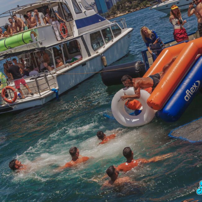 A group of people enjoy a party on a boat. One person slides down an inflatable slide into the water, where several others are swimming. The scene is lively, with sunshine and a city skyline in the background.