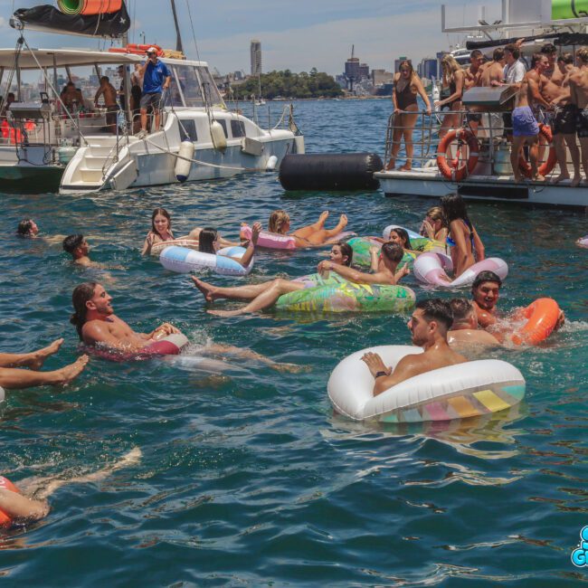 A group of people in colorful swimsuits float on inflatable tubes in the water near two boats, while others socialize on the boat decks under a sunny sky. The scene looks lively and festive.
