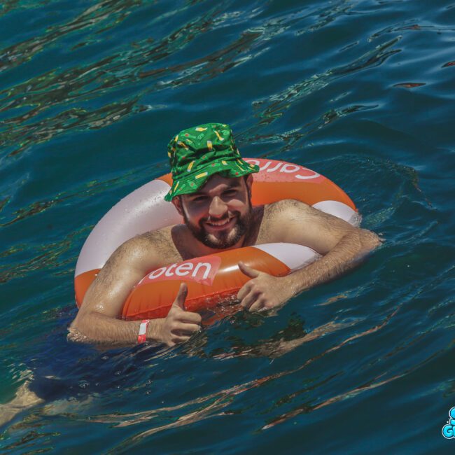 A man wearing a green bucket hat and wristband floats in blue water using an orange and white inflatable ring. He smiles and gives two thumbs up. Logos are visible at the bottom right corner of the image.