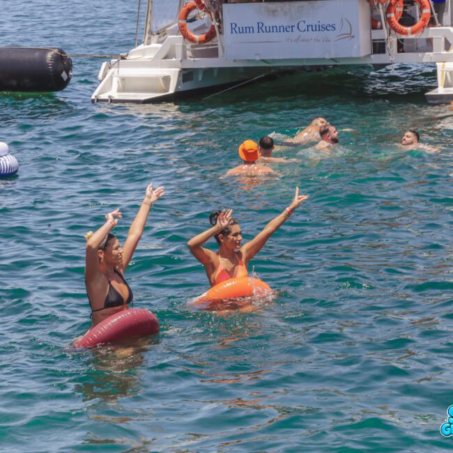 Two women in swim rings raise their arms and smile in the ocean near a boat labeled “Rum Runner Cruises,” surrounded by other people swimming and relaxing in the water on a sunny day.