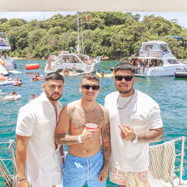 Three men posing on a boat, smiling at the camera during a lively party. Many people are seen swimming and socializing on surrounding boats in the sun near a green, tree-covered shore. The words "Sail Gustavo" are visible in the corner.