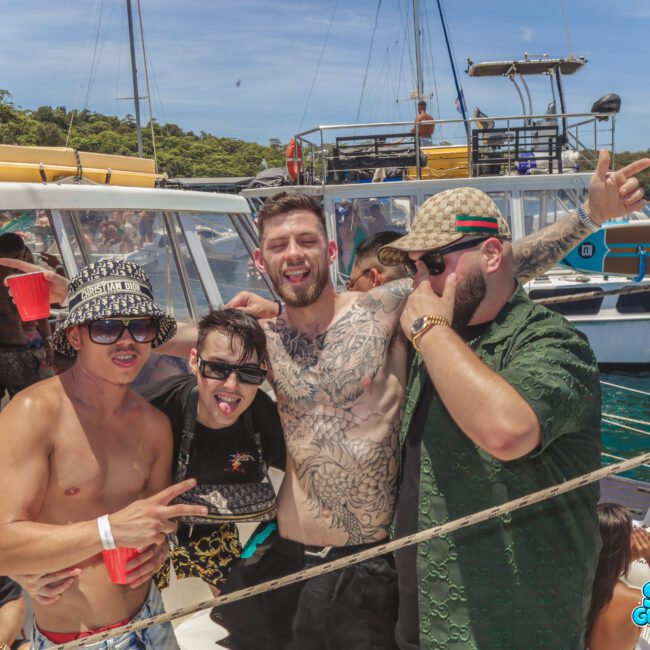 Four men pose together on a boat, smiling and enjoying a sunny day. Other people are in the background, some seated, with clear blue water and another boat visible behind them. The mood is festive and relaxed.