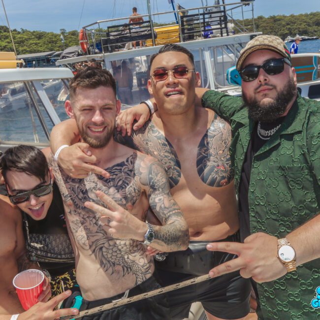 Five men pose together on a boat, smiling and making peace signs. Some are shirtless with visible tattoos, while others wear hats and sunglasses. The background shows water, trees, and more boats. It is sunny and festive.