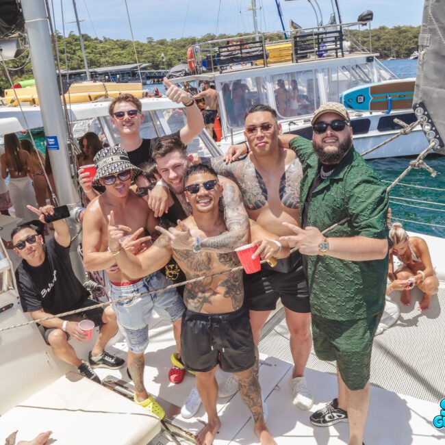 A group of men in swimwear pose and smile on the deck of a yacht at a lively boat party, with clear blue water, other boats, and trees in the background. Some hold drinks and make hand gestures. The logo "Sail Gustavo" is visible.