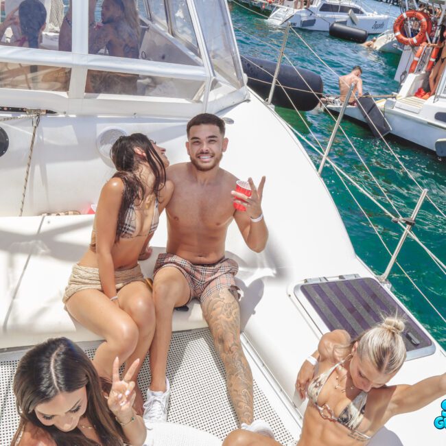 A group of young adults in swimsuits relax and pose for photos on a boat under bright sunlight, surrounded by blue water and other boats, with a "Sail Gustavo" logo in the corner.