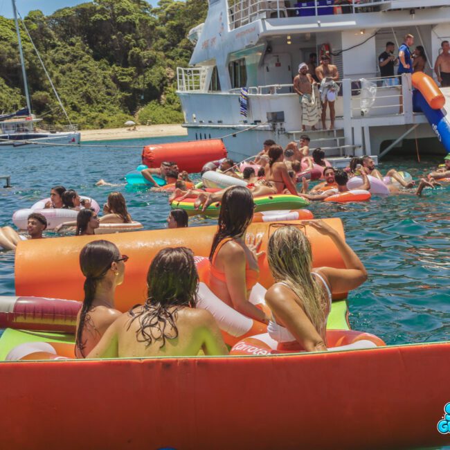 A group of people relax on colorful inflatables in the water near a large white boat, with others swimming and enjoying the sunny day; forested land is visible in the background.