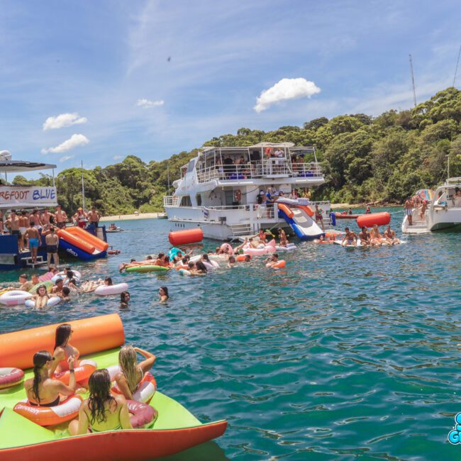 A large group of people enjoy a sunny day on colorful inflatables and boats in clear blue water, surrounded by green trees. Several boats are anchored close together, with people swimming and relaxing.