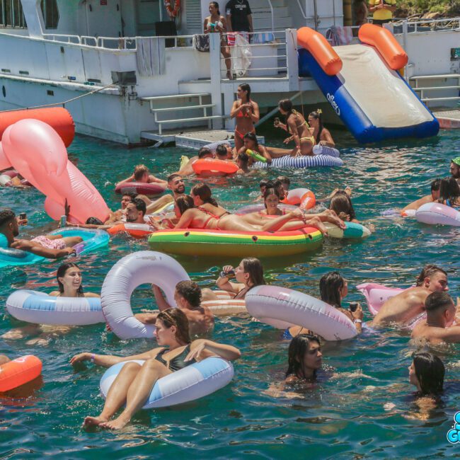 Dozens of people in swimsuits relax on colorful pool floats in the ocean by a white boat with slides. The water is clear and blue, and the scene is lively and festive under the sun.