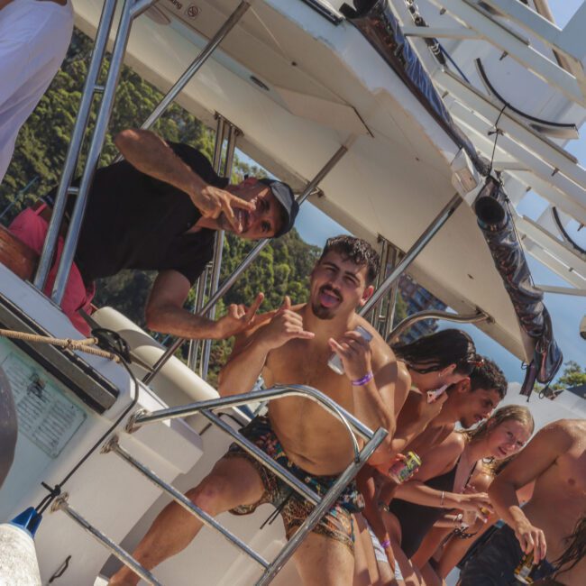 A group of young people in swimwear are having fun on a boat, smiling and posing for the camera near the water. Some are leaning over the railing, making hand signs, with trees and blue sky in the background.