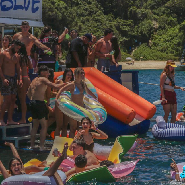 A group of young people enjoy a lively pool party on and around a boat, floating on inflatables in the water. The background shows lush greenery and another boat nearby. The mood is festive and energetic.
