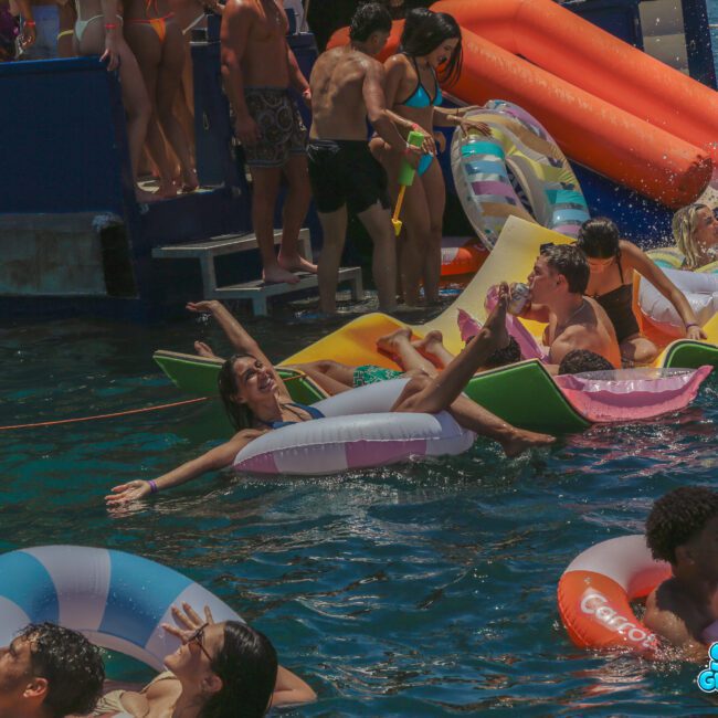 A group of young people relax and have fun on colorful pool floats in the water near a boat. Some are lounging, laughing, and socializing under the sun. The "Sail Gustavo" logo is visible in the bottom right corner.