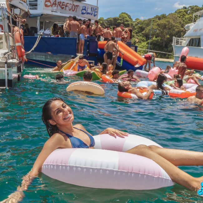 A woman in a blue bikini smiles while floating on a pink and white inflatable ring in the water, surrounded by other people on inflatables near docked boats during a lively outdoor party.