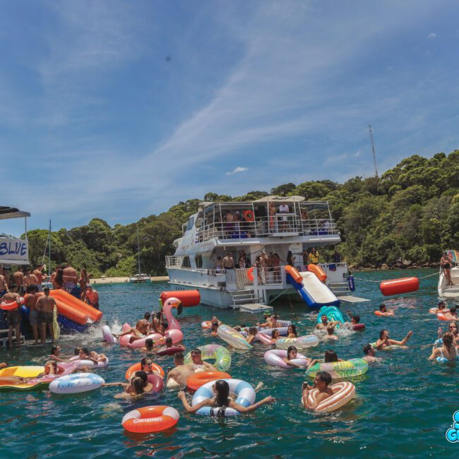 A lively scene of people relaxing on colorful inflatables in the water near boats, with lush green trees in the background and clear skies above. The atmosphere is festive and summery.