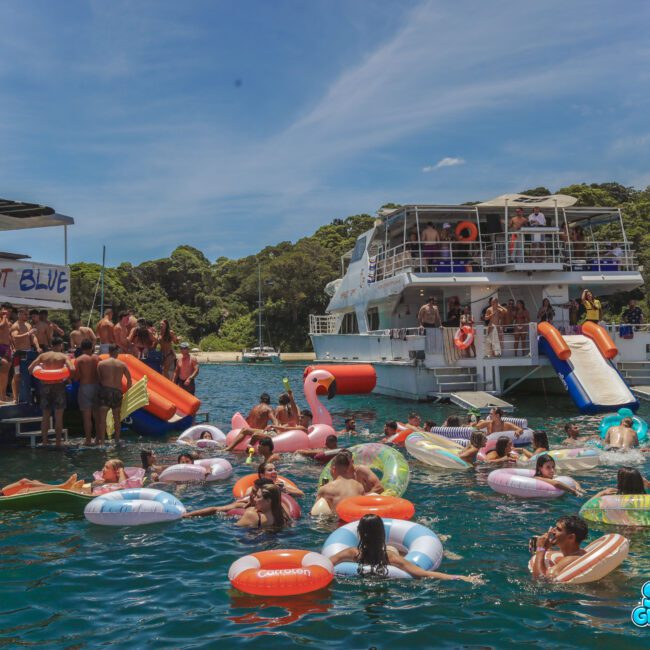 People enjoy a lively boat party, swimming and relaxing on colorful inflatables in clear blue water near two large boats; one has a waterslide, and the backdrop is lush green trees under a bright blue sky.
