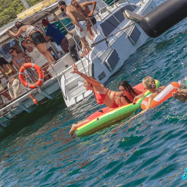 A group of people enjoy a sunny day on the water, some lounging on inflatable floats in the sea while others relax and take photos on a boat nearby. The scene is lively, with bright swimsuits and clear blue water.