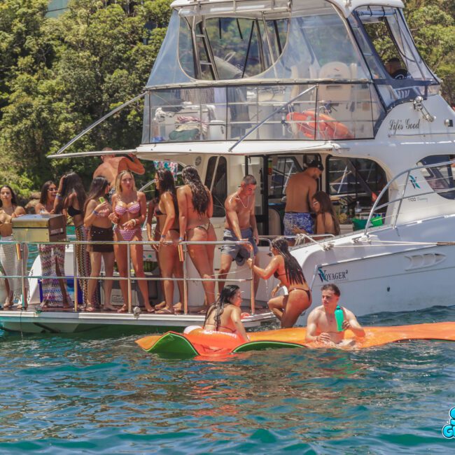 A group of people in swimsuits are gathered on a yacht and floating mats in blue-green water, surrounded by lush trees, enjoying a sunny day outdoors. The yacht has "Sail Gustavo" branding.