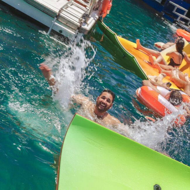 A man splashes joyfully into clear blue water while others relax on colorful floats nearby, next to a boat with people watching. "Sail Gustavo" logo is visible in the corner.