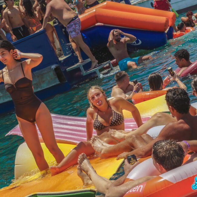 A group of young people relax on colorful inflatables and swim in clear blue water at a crowded pool party near a boat. Some are talking, posing, or sunbathing under bright sunlight. The "Sail Gustavo" logo is visible.