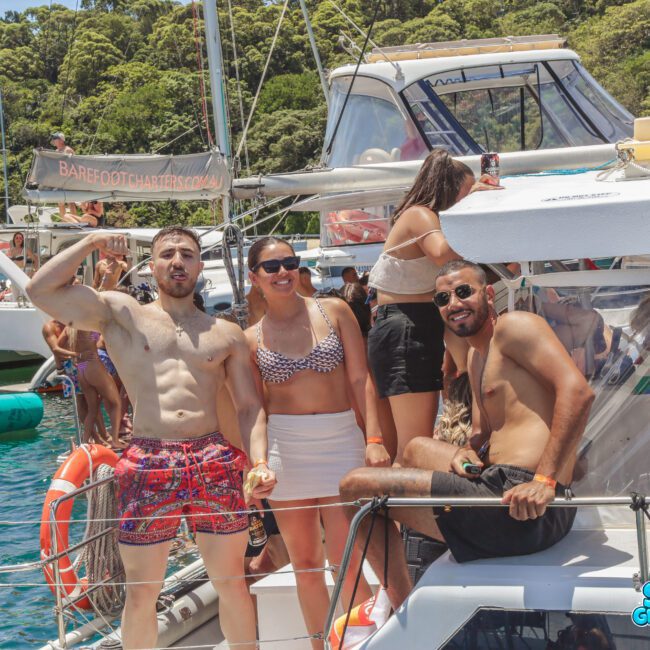 Three people in swimsuits smile and pose on a boat, surrounded by other boats and people on the water, with green trees in the background and a "Sail Gustavo" logo in the corner.