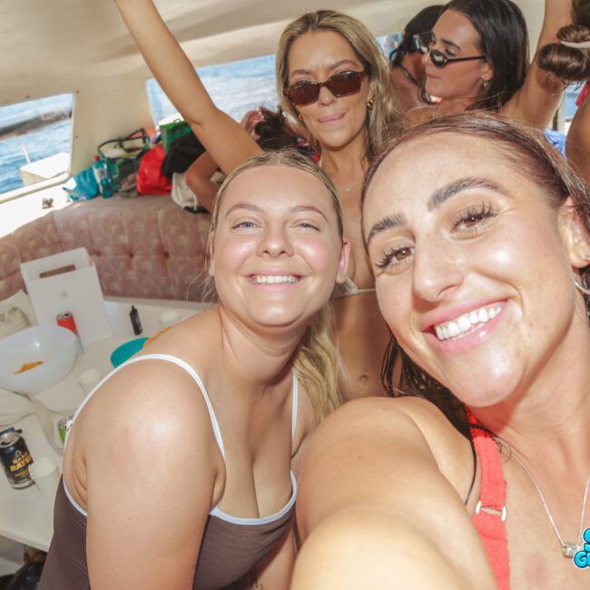 A group of women in swimsuits smile and take selfies while partying on a boat. The atmosphere is lively and festive, with drinks and snacks on a white table. Sunlight streams in through the boat windows.