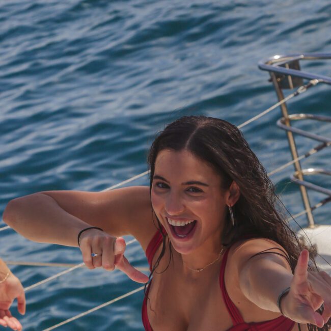 A smiling woman in a red bikini top poses playfully on a boat, pointing toward the camera with both hands. The ocean is visible in the background, and the boat railing is on the right. The "Sail Gustavo" logo appears at the bottom.
