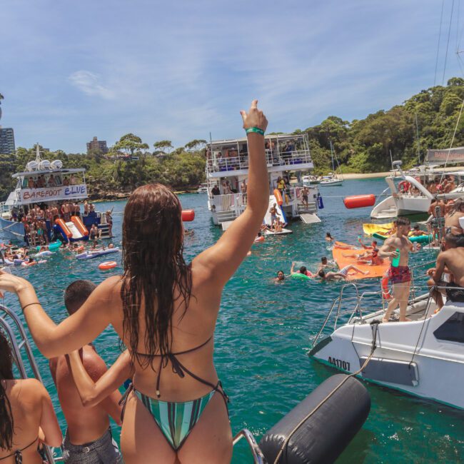 A woman in a bikini stands on a boat, raising her hand toward a lively scene of boats and people swimming and partying on the water under a sunny sky. The water is turquoise and several boats are anchored nearby.