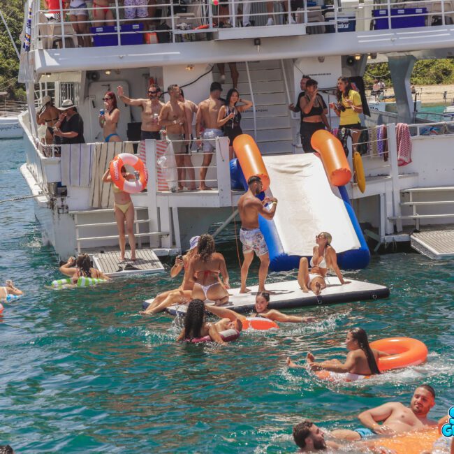 People enjoy swimming and relaxing on inflatables in the water near a boat with a slide. Others are on the deck, and the scene is lively with sun, fun, and colorful floaties. Sail Gustavo branding is visible.