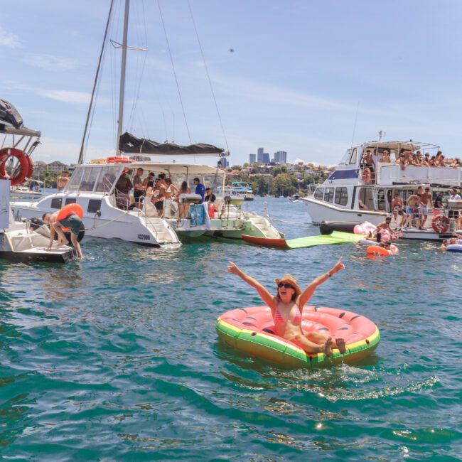 A woman on a watermelon-shaped float raises her arms while smiling in a lively harbor. People party on nearby boats under a sunny sky, with city buildings visible in the background. The water is clear and blue.