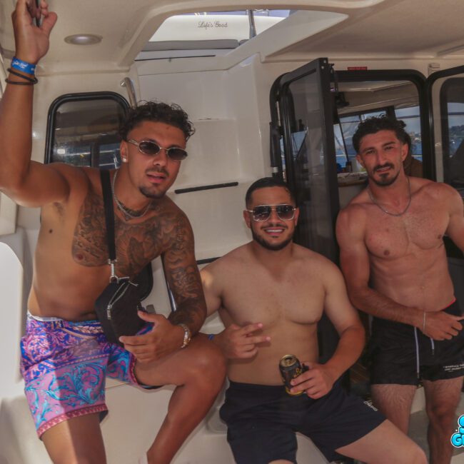 Three men in swimwear pose and smile on a boat. The man on the left has sunglasses and tattooed arms. The middle man is holding a can and wearing sunglasses. The right man is shirtless. Water and docks are visible in the background.