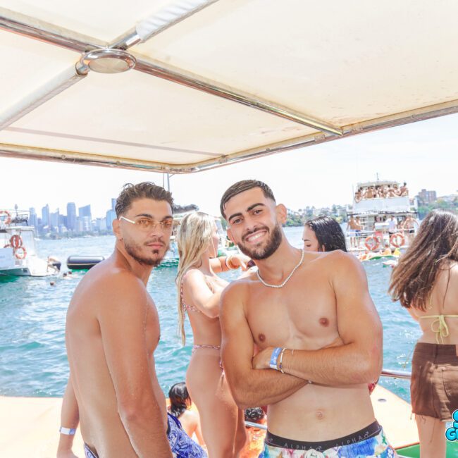 Two young men pose and smile on a boat at a lively party with many people in swimsuits, floating on the water and other boats in the background, with a city skyline visible in the distance.
