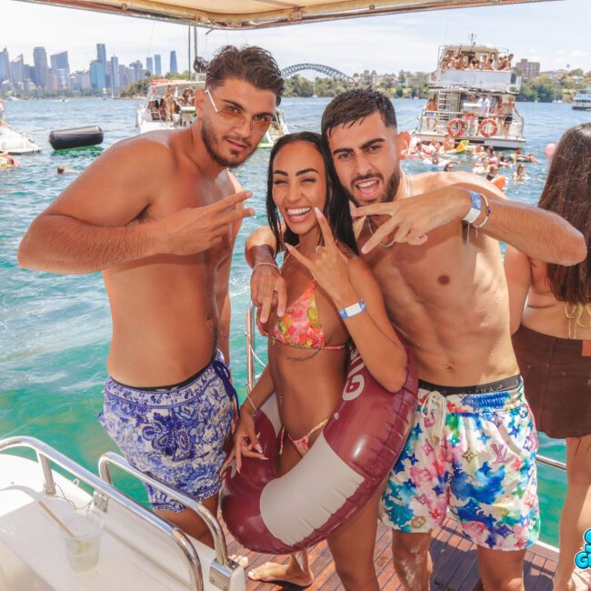 Three young adults in colorful swimwear pose and smile on a boat at a lively pool party, with people swimming and Sydney Harbour Bridge visible in the sunny background. The “Sail Gustavo” logo appears in the corner.